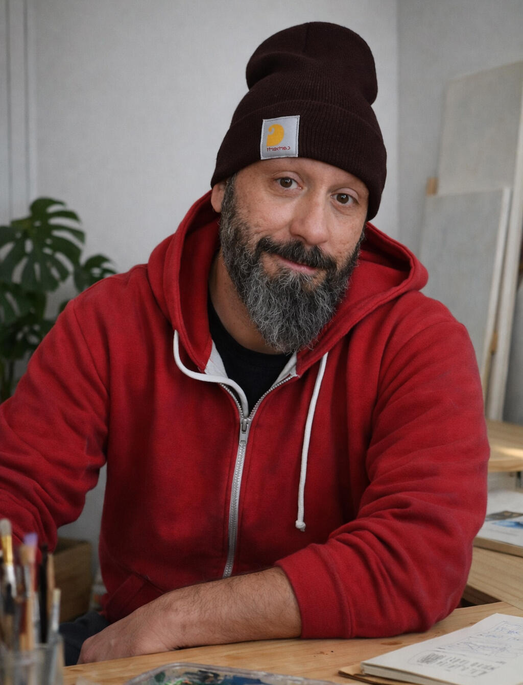 Isaac Núñez · Artista Automotriz Contemporáneo Fotografía de Isaac Núñez, artista contemporáneo especializado en la pintura de motor y vehículos clásicos, en su estudio creativo. El autor aparece mirando a cámara con una expresión cercana, con barba, vistiendo una sudadera roja y un gorro oscuro, rodea